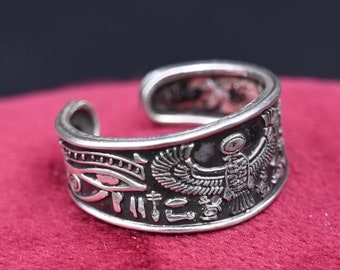 May include: Silver ring with an intricate design featuring an Egyptian eye and a winged creature. The ring is on a red velvet surface.