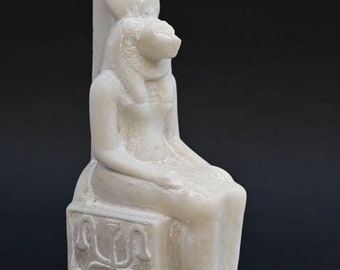 May include: White marble statue of an Egyptian goddess seated on a throne. The goddess has a lion head and wears a crown. The throne is decorated with hieroglyphs.