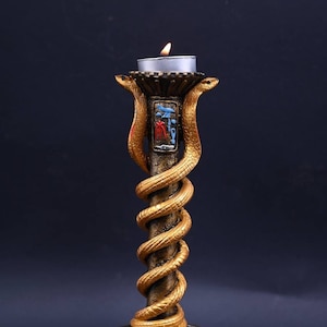 Może przedstawiać: Złoty świecznik z wężowym wzorem. Świecznik ma okrągłą podstawę i pojedynczy uchwyt na świecę na górze. Świecznik jest zapalony małą białą świecą.