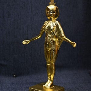 Gold leaf plated stone statue of the Egyptian goddess Neith standing with arms extended, wearing an elaborate headdress, photographed on a dark blue fabric background.