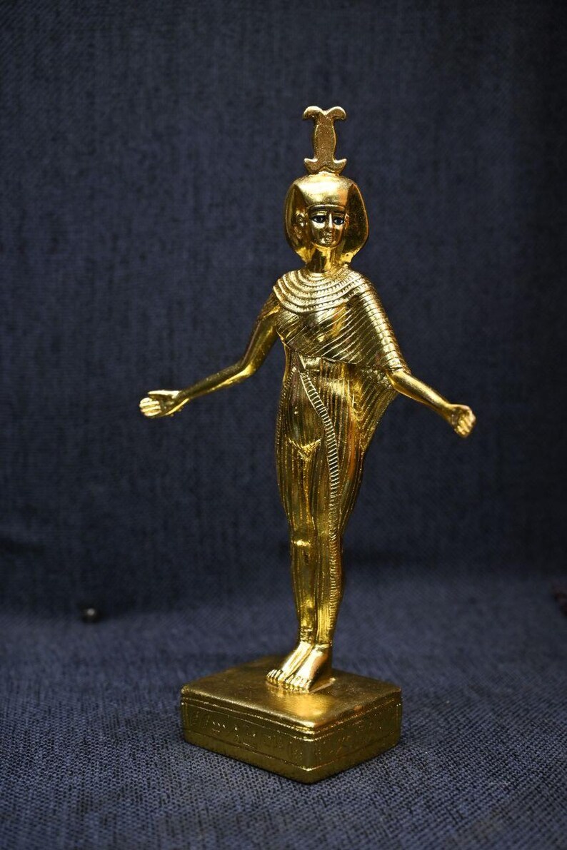 Gold leaf plated stone statue of the Egyptian goddess Neith standing with arms extended, wearing an elaborate headdress, photographed on a dark blue fabric background.