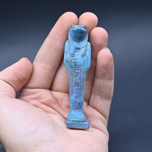 Puede incluir: Una figura ushabti egipcia de cer&aacute;mica vidriada azul y verde del dios Heqet, representado como una rana con brazos y piernas humanas. La figura est&aacute; de pie con jerogl&iacute;ficos inscritos en la parte delantera.