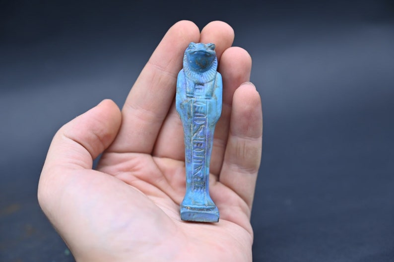 Puede incluir: Una figura ushabti egipcia de cer&aacute;mica vidriada azul y verde del dios Heqet, representado como una rana con brazos y piernas humanas. La figura est&aacute; de pie con jerogl&iacute;ficos inscritos en la parte delantera.