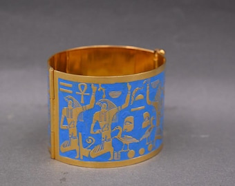 May include: Gold-coloured cuff bracelet with a blue enamel band featuring ancient Egyptian hieroglyphs. The bracelet has a hinged design and is likely made of metal. The blue enamel contrasts with the gold, creating a striking visual.