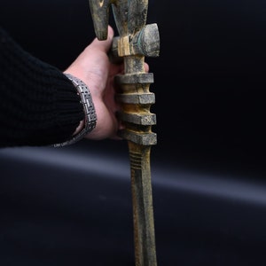 Puede incluir: Un bast&oacute;n de madera con una cabeza de p&aacute;jaro tallada en la parte superior. El bast&oacute;n est&aacute; pintado en tonos de negro, verde y marr&oacute;n. La cabeza de p&aacute;jaro tiene un ojo azul y un pico rojo.