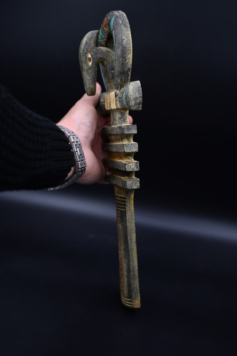 Puede incluir: Un bast&oacute;n de madera con una cabeza de p&aacute;jaro tallada en la parte superior. El bast&oacute;n est&aacute; pintado en tonos de negro, verde y marr&oacute;n. La cabeza de p&aacute;jaro tiene un ojo azul y un pico rojo.