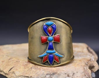 May include: Wide brass cuff bracelet featuring an Ankh symbol with red, blue, and turquoise enamel accents. The bracelet has a textured surface and is displayed on a stone.