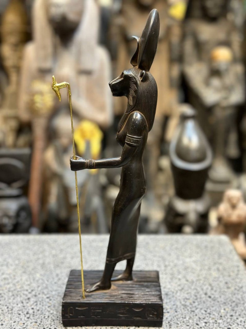 May include: Black wooden statue of the Egyptian goddess Bastet, depicted as a woman with a cat's head, holding a golden staff. The statue is standing on a wooden base with hieroglyphics.