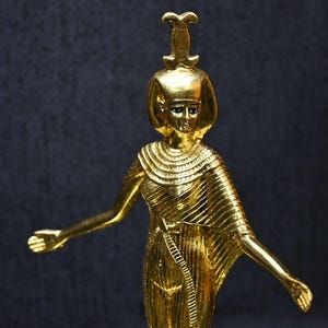 Gold leaf plated stone statue of the Egyptian goddess Neith standing with arms extended, wearing an elaborate headdress, photographed on a dark blue fabric background.