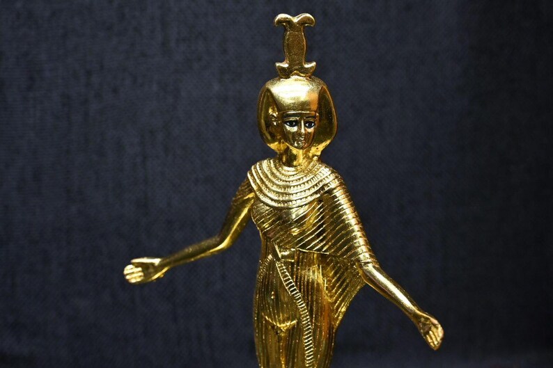 Gold leaf plated stone statue of the Egyptian goddess Neith standing with arms extended, wearing an elaborate headdress, photographed on a dark blue fabric background.