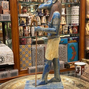 May include: A blue and gold statue of an Egyptian god with a jackal head, holding a staff. The statue is standing on a base with hieroglyphics.