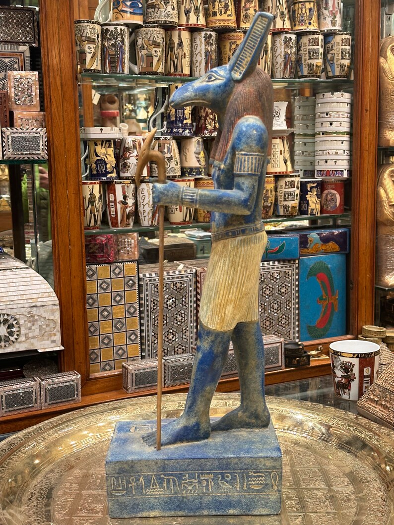 May include: A blue and gold statue of an Egyptian god with a jackal head, holding a staff. The statue is standing on a base with hieroglyphics.