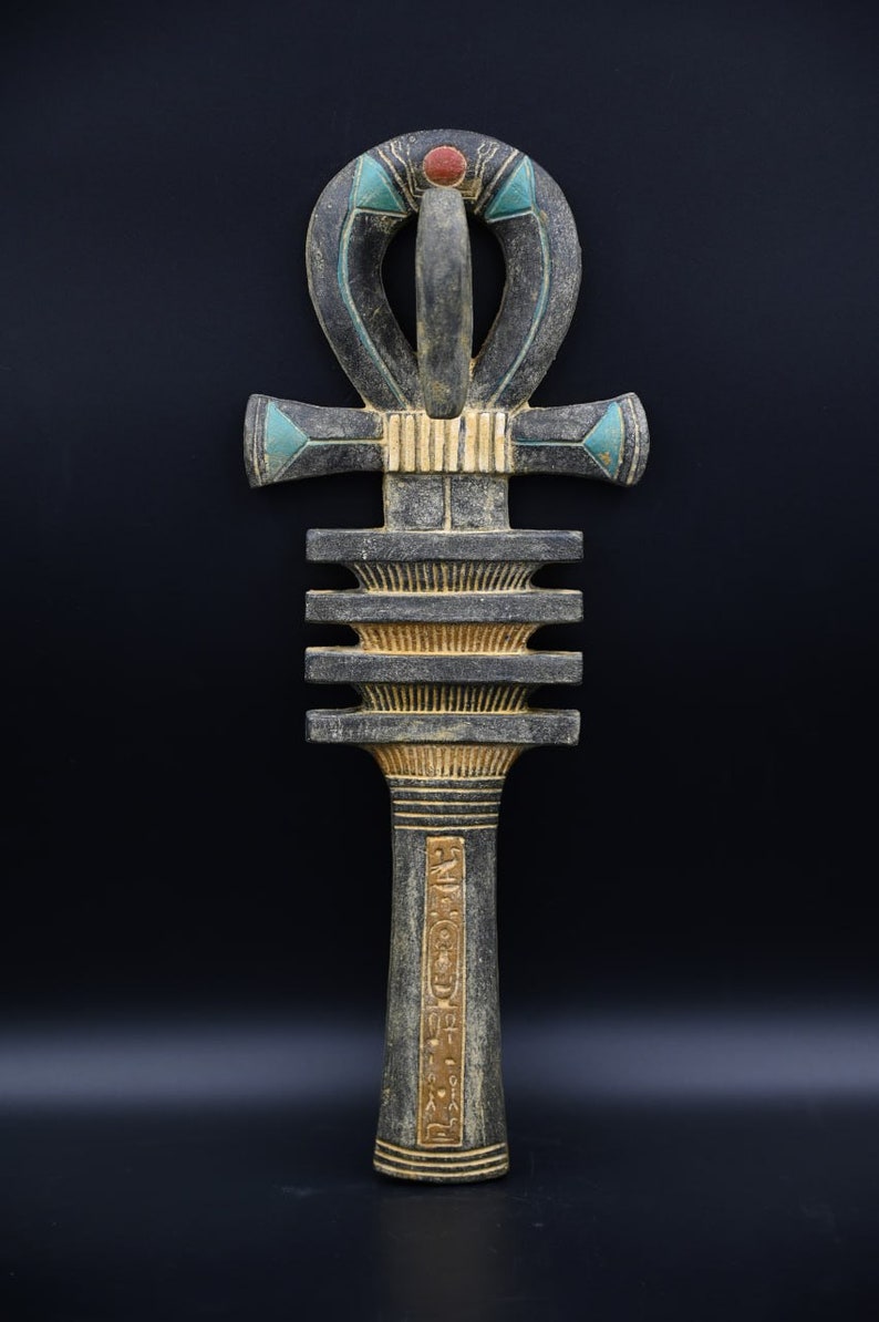 Puede incluir: Un Ankh de madera, un antiguo s&iacute;mbolo egipcio de la vida, con un acabado pintado en negro, dorado y azul. El Ankh est&aacute; decorado con jerogl&iacute;ficos y tiene un punto rojo en la parte superior.