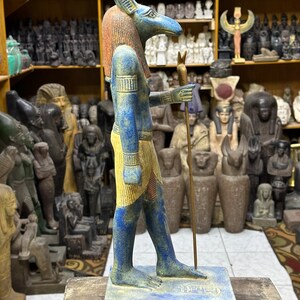 May include: A blue and gold painted statue of the Egyptian god Anubis, standing on a base with hieroglyphics. The statue is holding a staff in its right hand.