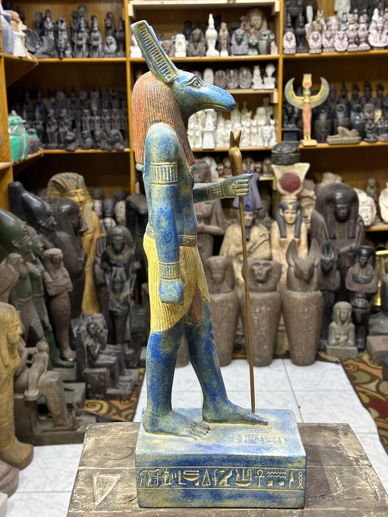 May include: A blue and gold painted statue of the Egyptian god Anubis, standing on a base with hieroglyphics. The statue is holding a staff in its right hand.