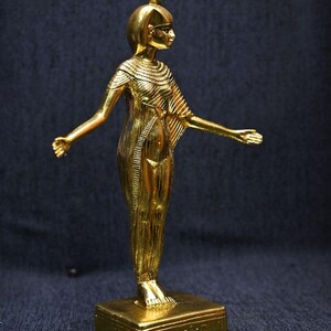 Gold leaf plated stone statue of the Egyptian goddess Neith standing with arms extended, wearing an elaborate headdress, photographed on a dark blue fabric background.