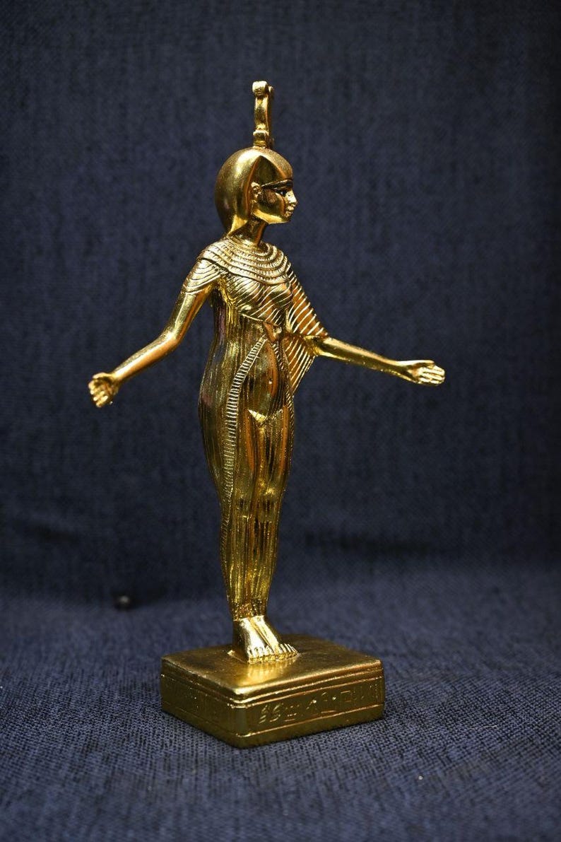 Gold leaf plated stone statue of the Egyptian goddess Neith standing with arms extended, wearing an elaborate headdress, photographed on a dark blue fabric background.