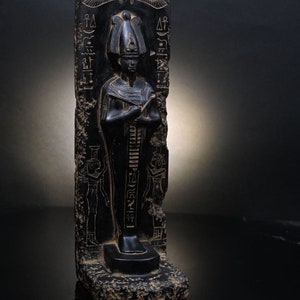 Puede incluir: Estatua de piedra negra de un fara&oacute;n egipcio con jerogl&iacute;ficos tallados en la piedra. El fara&oacute;n lleva una corona alta y una t&uacute;nica larga. La estatua est&aacute; de pie sobre una base con una peque&ntilde;a inscripci&oacute;n.
