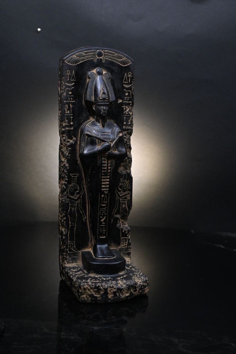 Puede incluir: Estatua de piedra negra de un fara&oacute;n egipcio con jerogl&iacute;ficos tallados en la piedra. El fara&oacute;n lleva una corona alta y una t&uacute;nica larga. La estatua est&aacute; de pie sobre una base con una peque&ntilde;a inscripci&oacute;n.