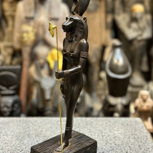 May include: Black statue of the Egyptian goddess Bastet, depicted as a woman with a lion head, holding a staff with a golden top. The statue is standing on a wooden base with hieroglyphs.