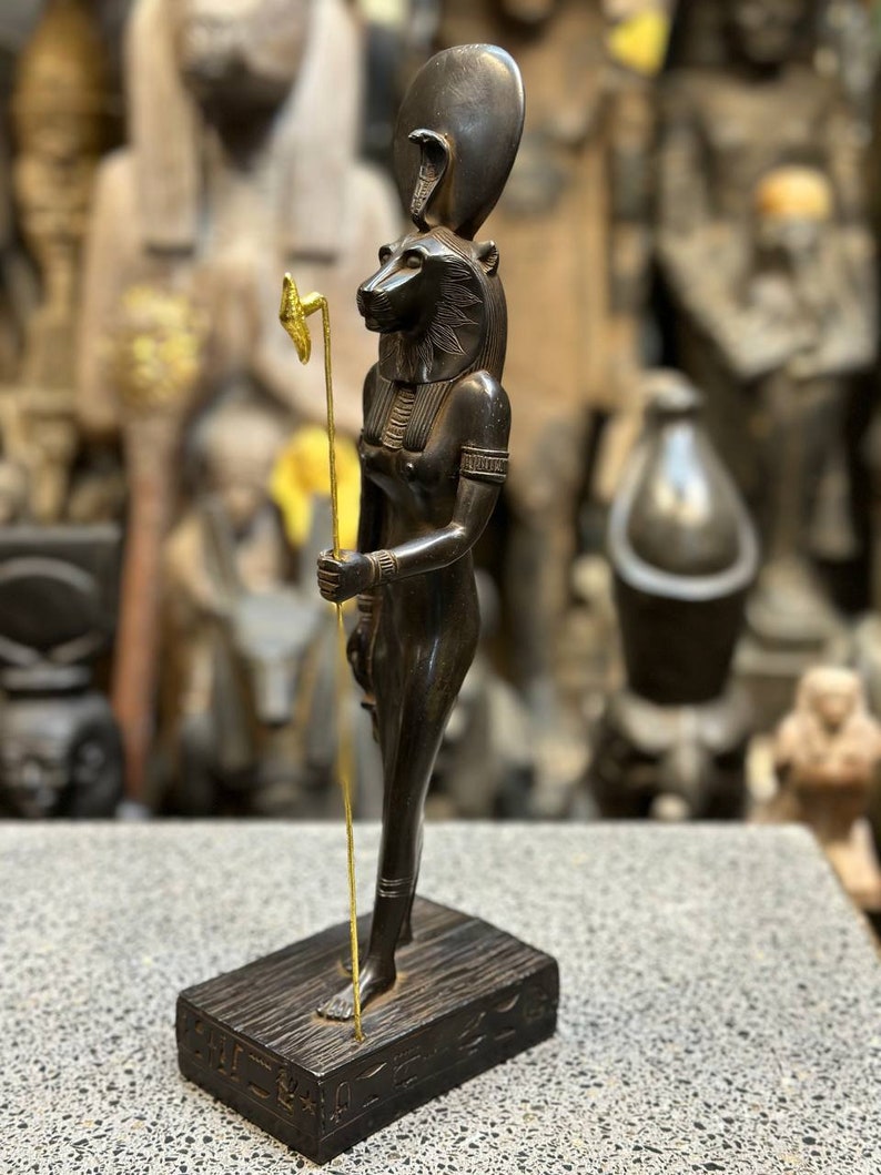May include: Black statue of the Egyptian goddess Bastet, depicted as a woman with a lion head, holding a staff with a golden top. The statue is standing on a wooden base with hieroglyphs.