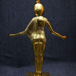 Gold leaf plated stone statue of the Egyptian goddess Neith standing with arms extended, wearing an elaborate headdress, photographed on a dark blue fabric background.