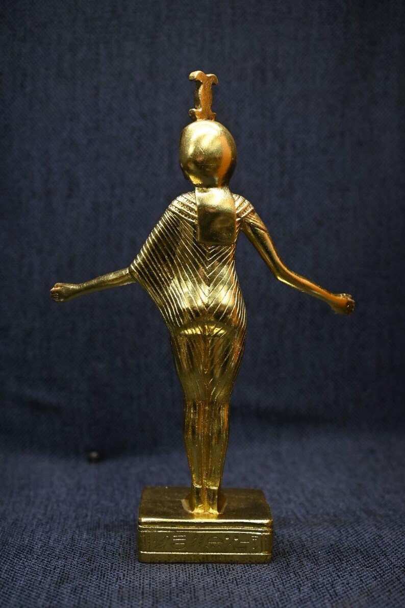 Gold leaf plated stone statue of the Egyptian goddess Neith standing with arms extended, wearing an elaborate headdress, photographed on a dark blue fabric background.