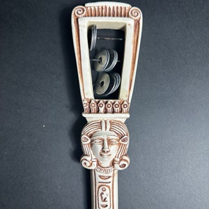 May include: White ceramic musical instrument with a carved female face and hieroglyphics. The instrument has a frame with metal discs that make a sound when shaken.