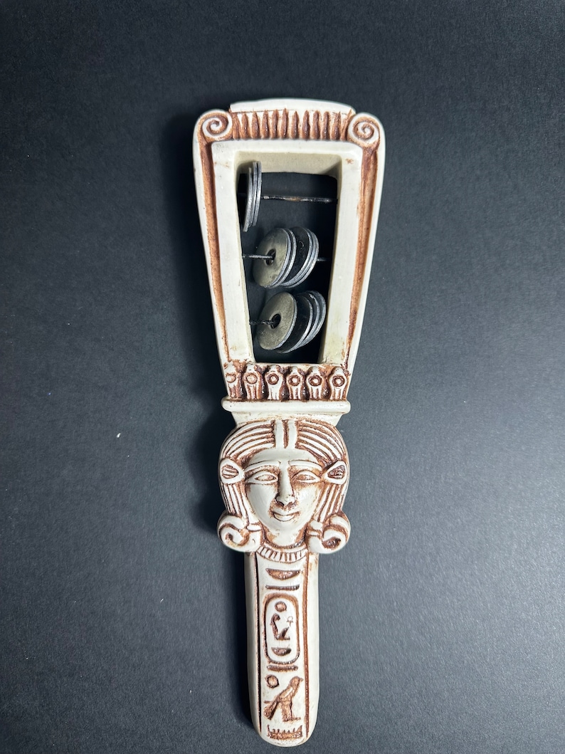 May include: White ceramic musical instrument with a carved female face and hieroglyphics. The instrument has a frame with metal discs that make a sound when shaken.