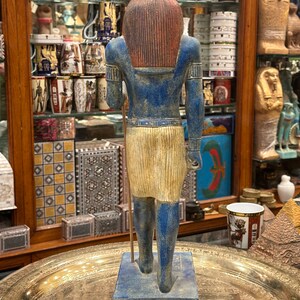 May include: A blue and gold painted statue of an Egyptian god with a jackal head. The statue is standing on a blue base and is holding a staff in its right hand.