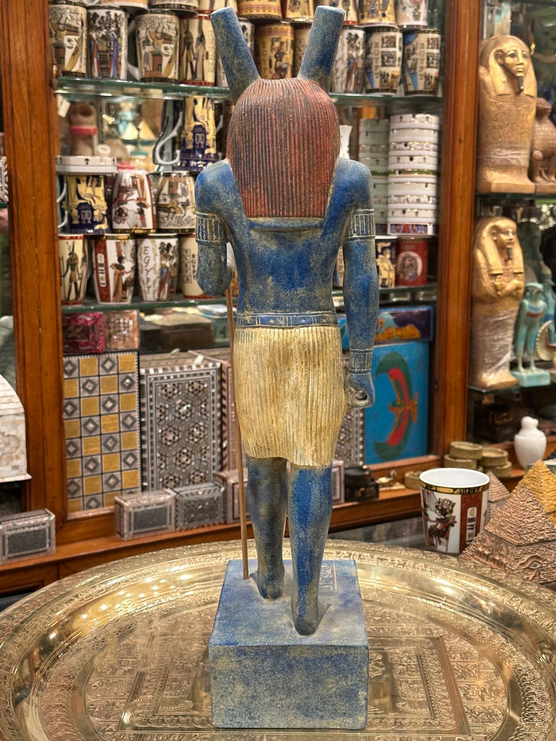 May include: A blue and gold painted statue of an Egyptian god with a jackal head. The statue is standing on a blue base and is holding a staff in its right hand.