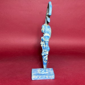 May include: A blue ceramic statue of an Egyptian goddess with a headdress and a cobra on her arm. The statue is standing on a base with hieroglyphics.