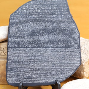 Könnte beinhalten: Eine schwarze Steinreplik des Rosetta-Steins, ein berühmtes Artefakt mit alten ägyptischen Hieroglyphen, Griechisch und Demotic-Schrift. Der Stein steht auf einem schwarzen Ständer und ein weißer Stein befindet sich im Vordergrund.
