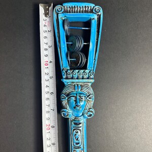 May include: A blue and black ceramic rattle shaped like an Egyptian goddess with a crown and a face. The rattle has a series of metal discs that make a sound when shaken.