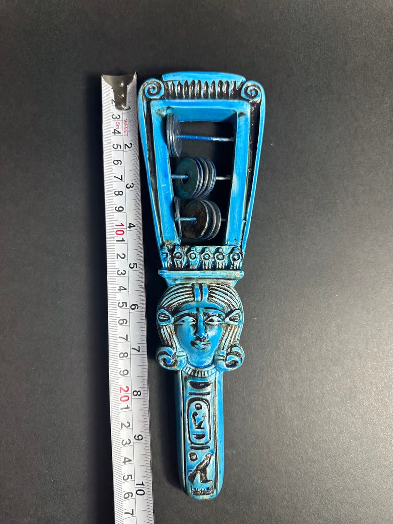 May include: A blue and black ceramic rattle shaped like an Egyptian goddess with a crown and a face. The rattle has a series of metal discs that make a sound when shaken.