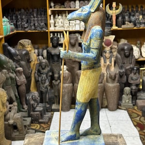 May include: A blue and gold statue of an Egyptian god with a jackal head, holding a staff with a forked top. The statue is standing on a base with hieroglyphics.