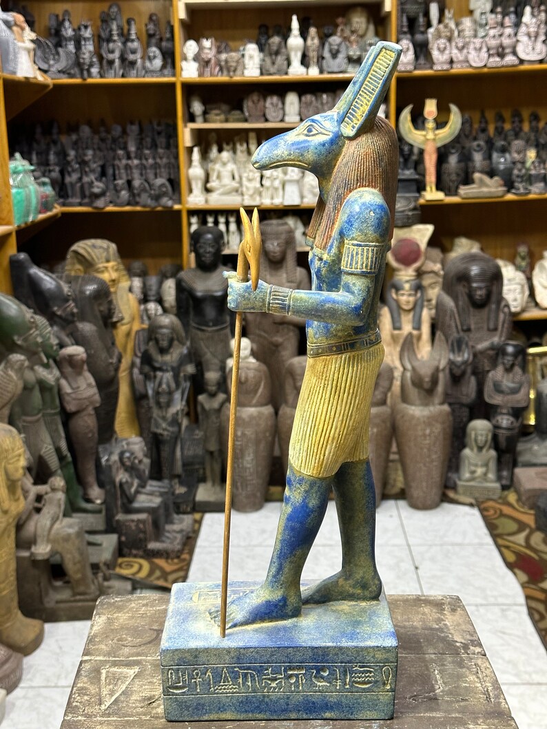 May include: A blue and gold statue of an Egyptian god with a jackal head, holding a staff with a forked top. The statue is standing on a base with hieroglyphics.