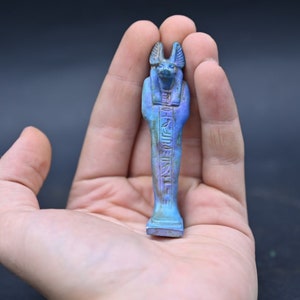 Puede incluir: Una peque&ntilde;a figura azul de un dios egipcio con cabeza de chacal. La figura est&aacute; tallada con jerogl&iacute;ficos y tiene una cara detallada.