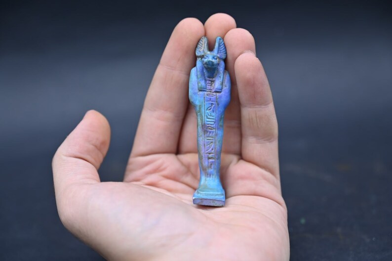 Puede incluir: Una peque&ntilde;a figura azul de un dios egipcio con cabeza de chacal. La figura est&aacute; tallada con jerogl&iacute;ficos y tiene una cara detallada.