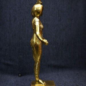 Gold leaf plated stone statue of the Egyptian goddess Neith standing with arms extended, wearing an elaborate headdress, photographed on a dark blue fabric background.