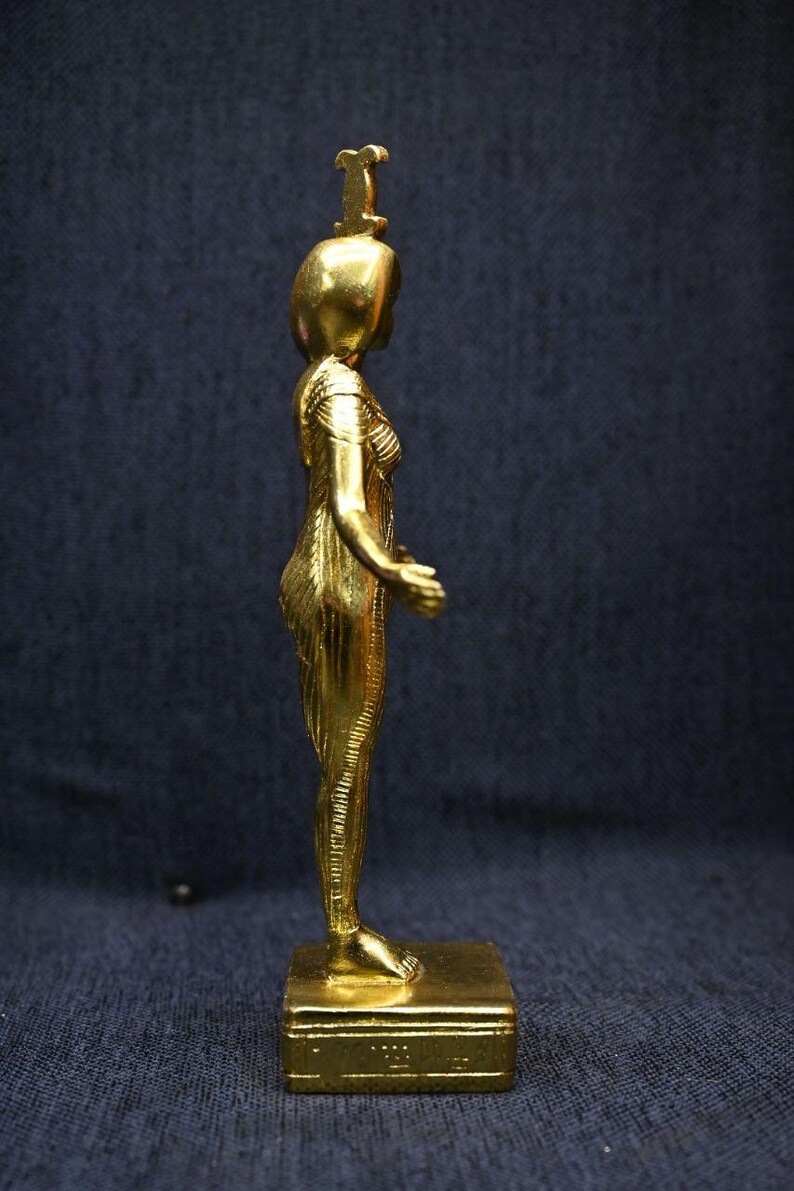 Gold leaf plated stone statue of the Egyptian goddess Neith standing with arms extended, wearing an elaborate headdress, photographed on a dark blue fabric background.