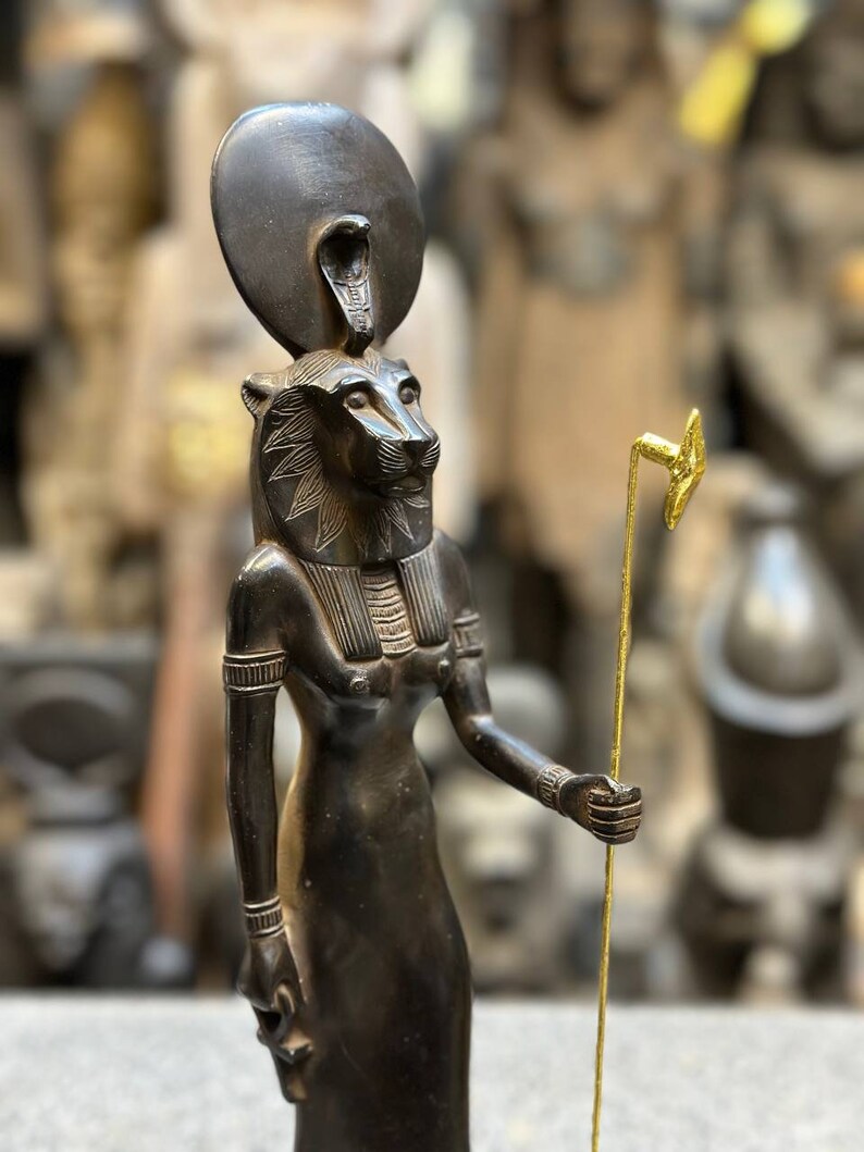 May include: Black statue of the Egyptian goddess Sekhmet, depicted as a lioness with a sun disk and cobra on her head. She holds a golden staff in her right hand.
