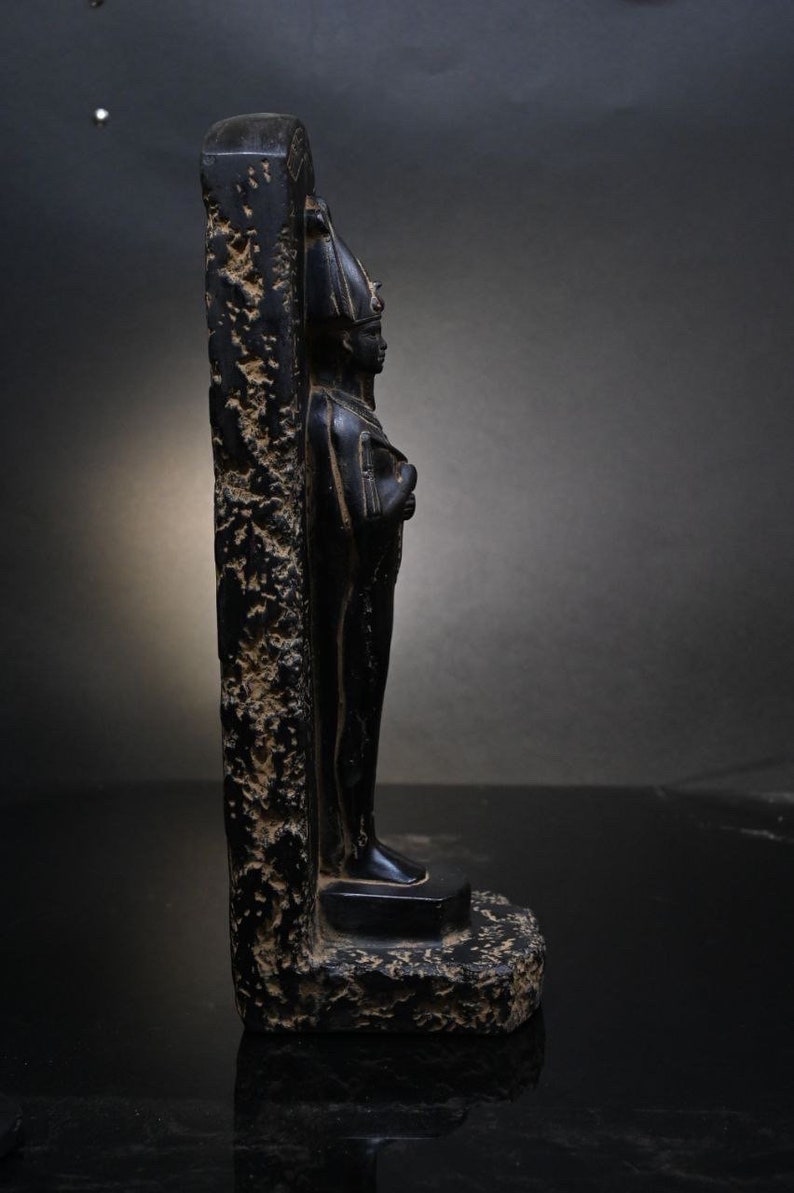 Puede incluir: Estatua de piedra negra de un dios o fara&oacute;n egipcio de pie en un bloque de piedra tallado. La estatua es detallada y tiene un aspecto desgastado.