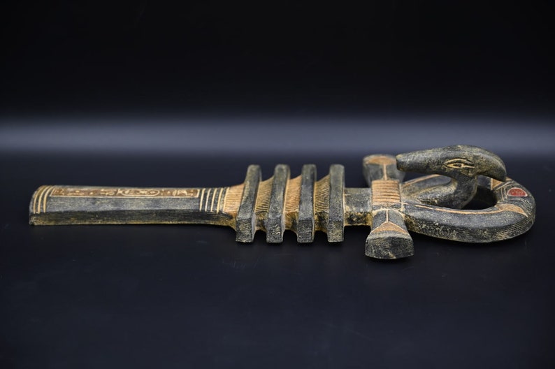 以下が含まれることがあります： 古代エジプトの生命の象徴である、彫刻された木製のアンク。アンクは主に濃い茶色で、薄い茶色と金のアクセントがあります。上部には、赤いアクセントが付いた様式化されたヘビの頭があります。