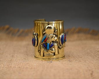 May include: A gold-coloured metal cuff bracelet with an Egyptian-style design. The cuff features a raised image of a pharaoh with a blue and red headdress and a blue and red collar. The cuff is decorated with blue and red gemstones.