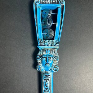 May include: A blue and black wooden rattle with a carved image of an Egyptian goddess and a series of metal discs that make a rattling sound when shaken.