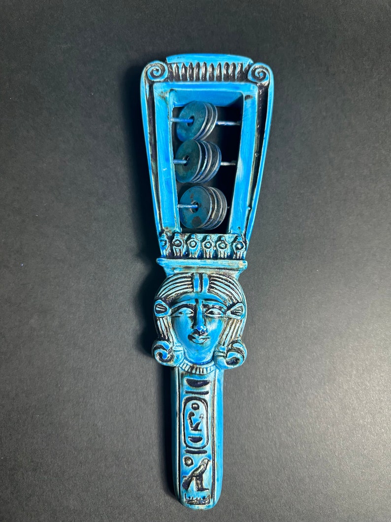 May include: A blue and black wooden rattle with a carved image of an Egyptian goddess and a series of metal discs that make a rattling sound when shaken.