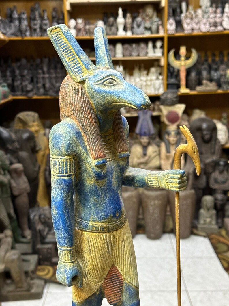 May include: A blue and gold statue of the Egyptian god Anubis, depicted as a jackal-headed human with a staff. The statue is standing on a wooden base.