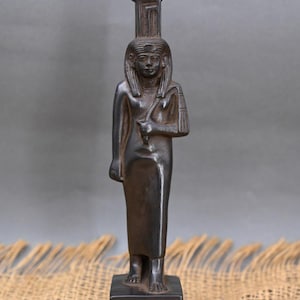 Peut inclure: Une statue noire d'une déesse égyptienne debout sur une base carrée avec des hiéroglyphes. La déesse porte une longue robe et une couronne avec un sommet en forme de coupe haut et fin.