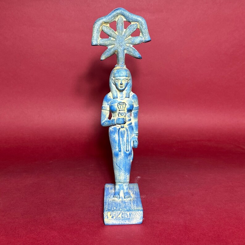 May include: A blue ceramic statue of an Egyptian goddess holding a staff with a sun symbol on top. The statue is standing on a rectangular base with hieroglyphs.